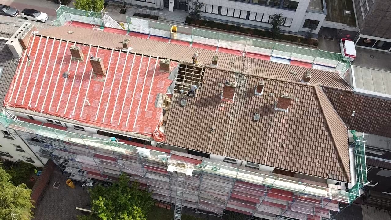 Luftaufnahme einer Dachsanierung: eine Dachhälfte bereits neu mit Ziegeln eingedeckt, die andere zeigt die freiliegende rote Abdichtungsbahn, Gerüst rundum am Gebäude.