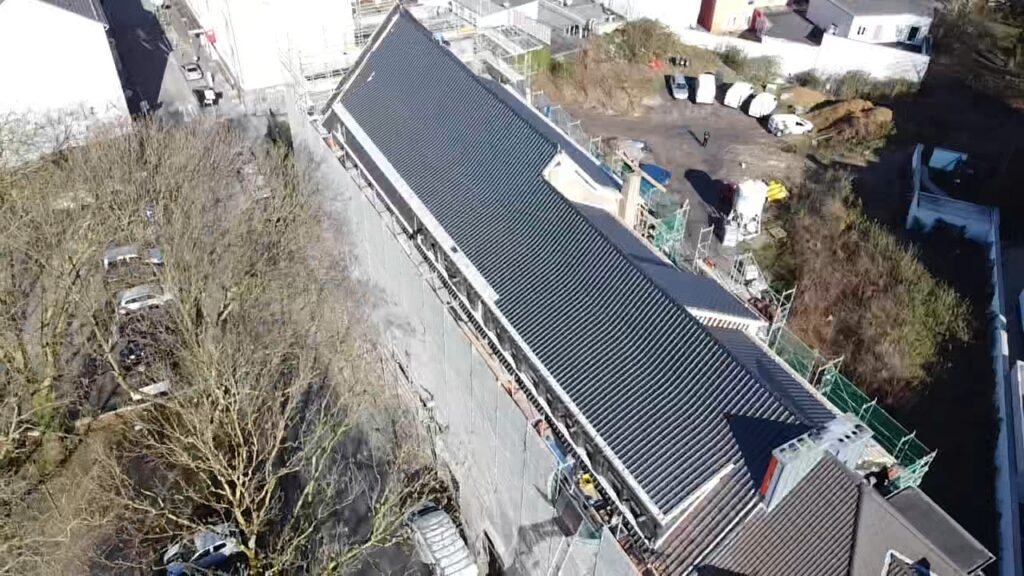Luftaufnahme eines Mehrfamilienhauses in Bochum während der Neueindeckung: eine Dachhälfte mit neuen anthrazitfarbenen Ziegeln, Gerüst entlang der Fassade.